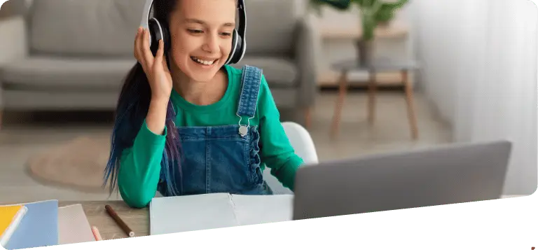 Jeune fille souriante en salopette verte, portant un casque et suivant un cours d'anglais en ligne depuis un ordinateur portable